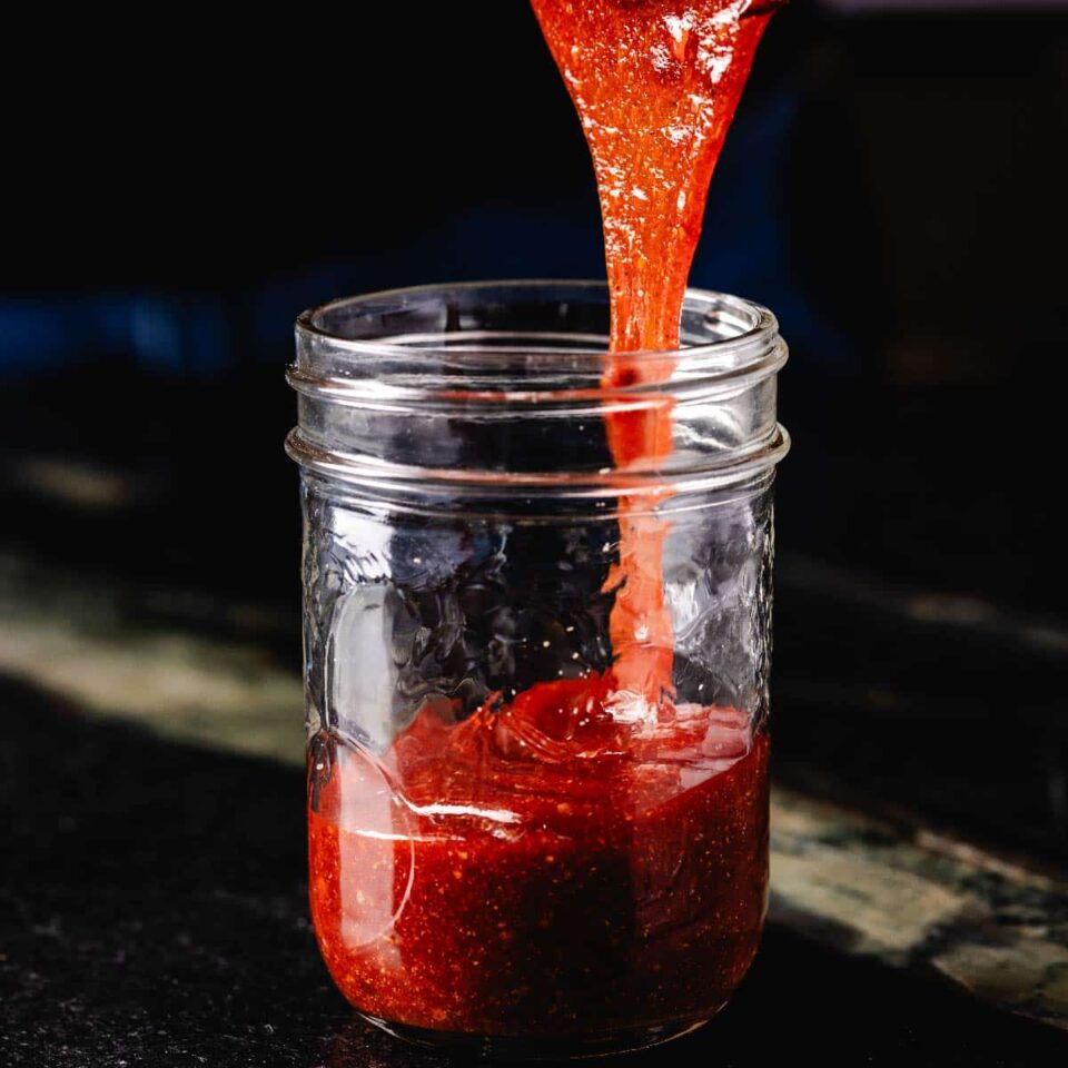 A glass jar with a red sauce being poured into it.
