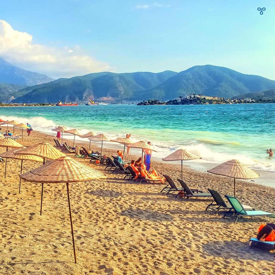 People sunbathing on Çalış beach with waves crashing. Mountains are in the background.