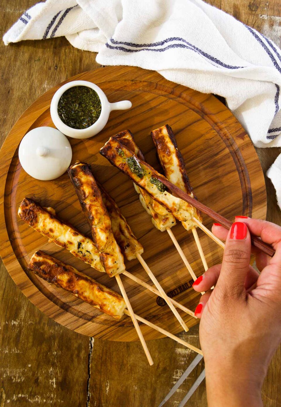 a hand drizzles chimichurri sauce over grilled queijo coalho on a wooden platter