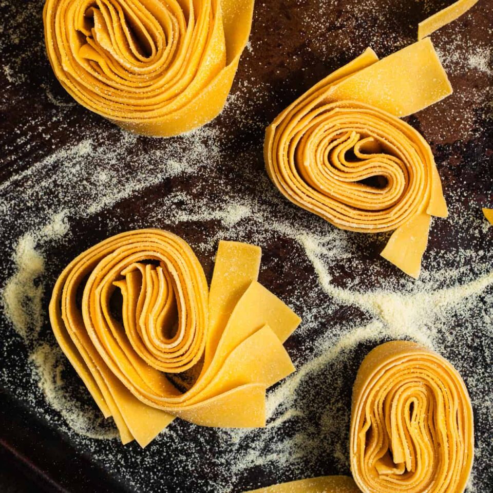 Nests of pappardelle pasta dusted in semolina flour on a baking sheet.