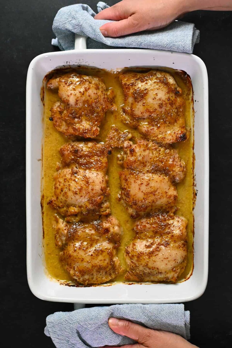 An overhead shot of someone holding a rectangular white baking dish filled with baked honey mustard chicken thighs.