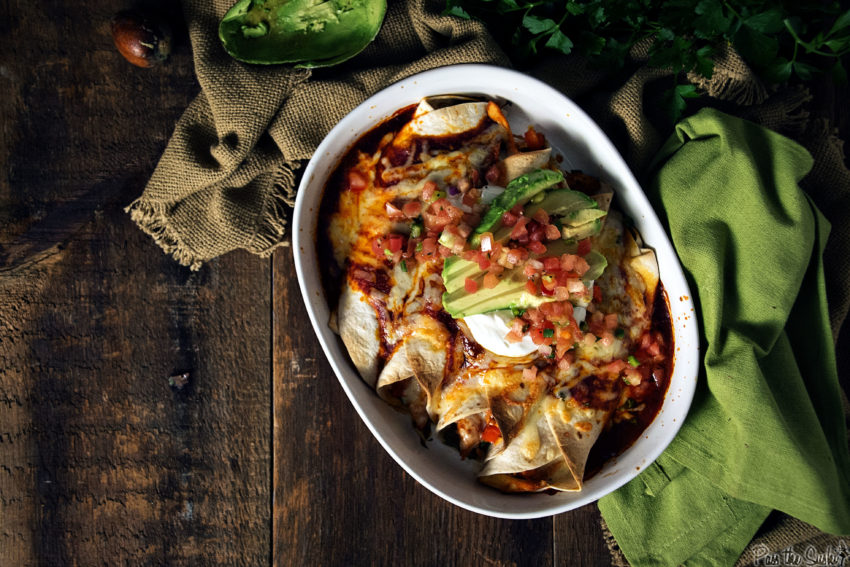 enchiladas with pico de gallo, sour cream and sliced avocado in a white dish on a wooden table with green linen